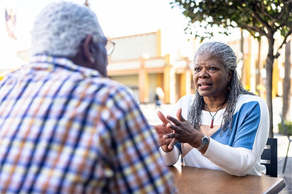 Black older couple talking together across a table outside. 