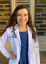 Student Courtney Bair in white coat and stethoscope standing in front of brick wall with hand on hip
