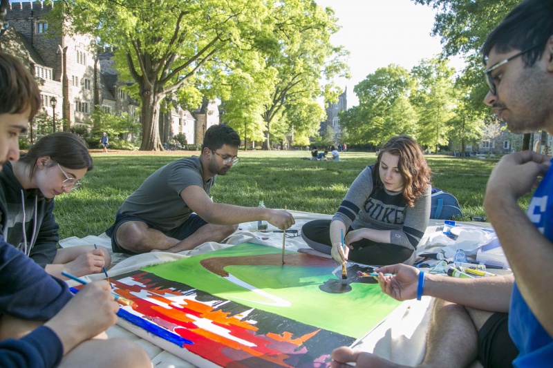 Students sitting on the Duke Quad around a canvass on the grass upon which they're painting a banner.