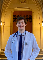 Student Sage Atkins in White Coat & Stethoscope in front of wooden door