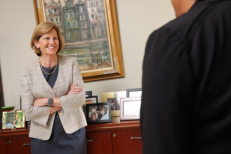 woman leans against desk speaking with another person