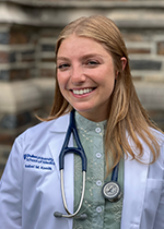 Student Isabel Koolik wearing white coat and stethoscope in front of brick wall