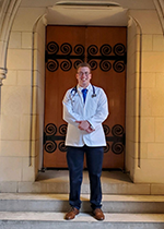 Student Judah Kreinbrook wearing white coat and stethoscope standing on steps in front of wooden door