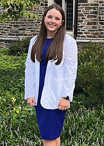 Student Lexy Markunas wearing white coat and blue dress standing in front of brick wall and greenery