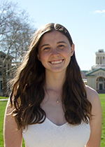 Headshot of student Katie Parsons wearing white shirt in front of blue sky and grass with brown hair over both shoulders