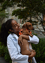 Student Janel Ramkalawan wearing white coat, holding small brown dog in front of trees