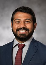 Headshot of student Jay Rathinavelu wearing navy suit and crimson tie in front of grey background