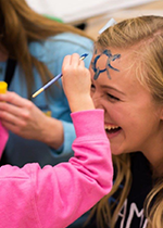 Student Claire Washabaugh getting blue face paint put on her face by small child's arm wearing pink sweatshirt