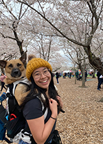 Student Marilyn Yamamoto wearing mustard coloured beanie holding a brown dog in backpack in front of bare trees