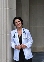 Student Katelyn Young wearing white cost and stethoscope leaning against pillars holding edges of her white coat