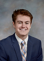 Student Lucas Collins smiling at camera wearing navy blazer and argyle tie in front of grey backdrop