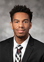 headshot of student ronald Harris wearing black blazer over white shirt and checkered tie in front of grey backdrop