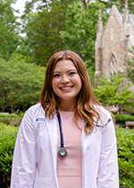 Student Taylor Kohlmann smiling at th ecamera wearing white coat and stethoscope over light pink dress in front of greenery and a brick building