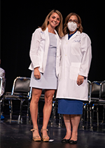 Student Sally Kuehn wearing white coat over blue dress on stage with a masked woman also wearing a white coat