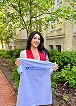 Student Hana Shafique holding grey colored Duke T shirt in front of her, smiling at camera in front of a tree
