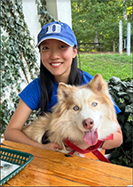 Student Avivah Wang sitting down behind a table wearing blue Duke cap and Blue T shirt, holding a light brown and white dog on her lap that is looking at camera with its tongue out