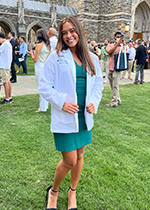 Student Rebecca Zasloff smiling at camera wearing white coat over green dress holding seams of coat standing on green grass in front of brick building