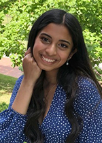 Neha Majety smiling at camera wearing blue polka dot shirt, curled hair in front of shoulders with right hand fisted resting under chin in front of greenery
