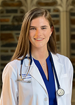 Jazlyn Seidel smiling at camera wearing white coat and stethoscope over blue dress in front of stone wall, straight hair is down behind her shoulders
