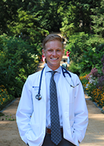 Trevor Sytsma smiling at camera wearing white coat and stethoscope over white business shirt and tie with hands in pocket, standing on a gravel track in park