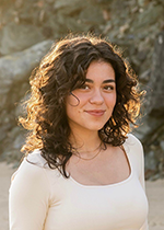 Raquel Garcia smiling with closed mouth at camera, brown curly shoulder length hair, wearing white long sleeve shirt standing on beach in front of large rock