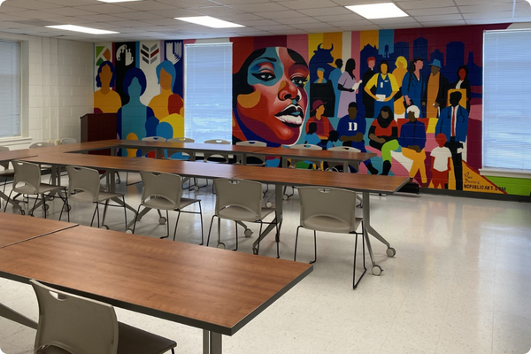A classroom with long tables and chairs and a mural depicting a diverse group of people.