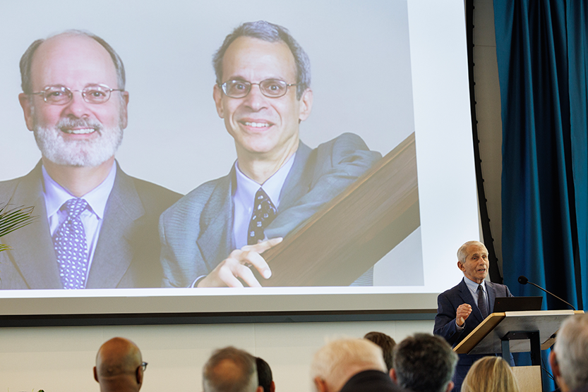 Dr. Fauci speaking in front of an image of Drs. Haynes and Letvin