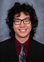 Headshot of Matthew Li smiling at camera, in front of blue backdrop, long black curly hair haloing head, wearing black framed glasses, red shirt and silver tie with black suit