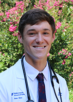 Headshot of student smiling at camera with dark brown hair wearing blue floral tie, white coat, and stethoscope in front of floral tree