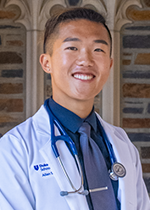 Headshot of student smiling at camera with black hair wearing blue tie, white coat, and stethoscope in front of brick wall