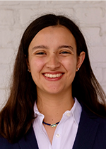 Headshot of student smiling at camera with dark brown straight hair wearing black blazer over white collared shirt in front of white brick wall