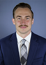 headshot of student with moustache wearing navy suit with black and grey checked tie in front of blue backdrop