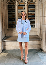 Blonde student standing in front of ornate brick wall, wearing White Coat and Stethoscope over short white dress and tan wedge heels,