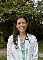 Brown haired student smiling at camera wearing white coat and stethoscope over green printed dress in front of tree
