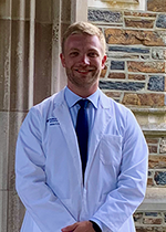 Student with blonde hair and stubble smiling at camera wearing white coat over navy tie and blue shirt, hands clasped in front of body standing in front of brick wall