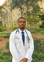 Student wearing white coat and stethoscope over white shirt and blue tie, hands clasped in front of body, standing in front of greenery and building