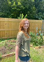 Red-haired student smiling, standing at angle from camera wearing denim shorts and green shirt, in front of fence and garden beds