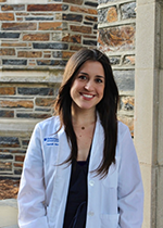 Student smiling at camera with brown straight hair falling in front of shoulders wearing white coat over black dress in front of brick wall