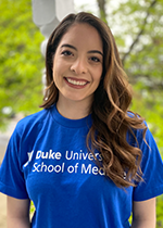 Student smiling at camera with brown curled hair over left shoulder, wearing lipstick, and Blue T-shirt that reads "Duke University School of Medicine"