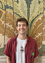 Brown haired student smiling wearing red button down open with white T-shirt underneath, standing in front of ornately tiled wiled