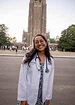 Brown haired Student smiling at camera wearing glasses, white coat and stethoscope standing on road in front of Duke Chapel
