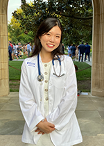 Black-haired students smiling at camera wearing white coat and stethoscope over cream dress with hands clasped in front of body