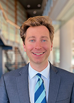 Headshot of Jack Finlay smiling at camera, short blonde hair, wearing charcoal suit with 2 toned blue striped tie, in front of blurred stairs in the background