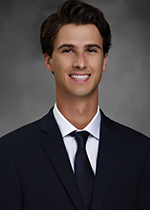 Headshot of Casey Gilles smiling at camera in front of grey backdrop wearing black suit and navy blue tie