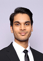 Headshot of Sunag Udupa wearing black suit and black tie slightly smiling at camera in front of grey backdrop