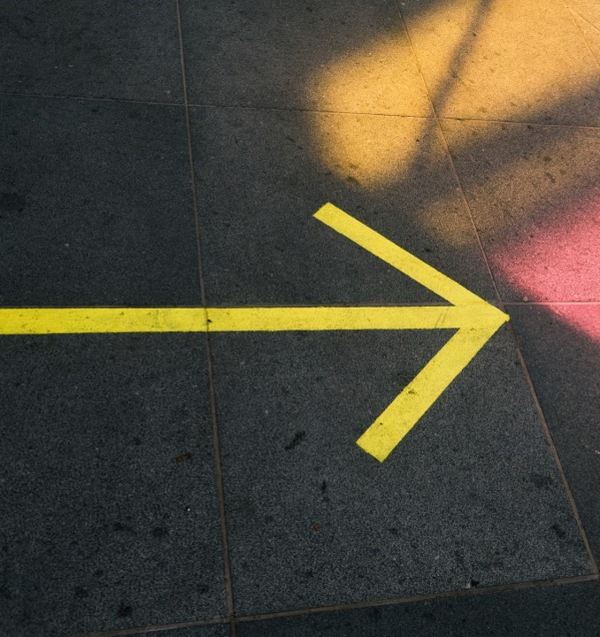 Image of a yellow arrow on the sidewalk pointing to the right, with sun shining on sidewalk through a window.