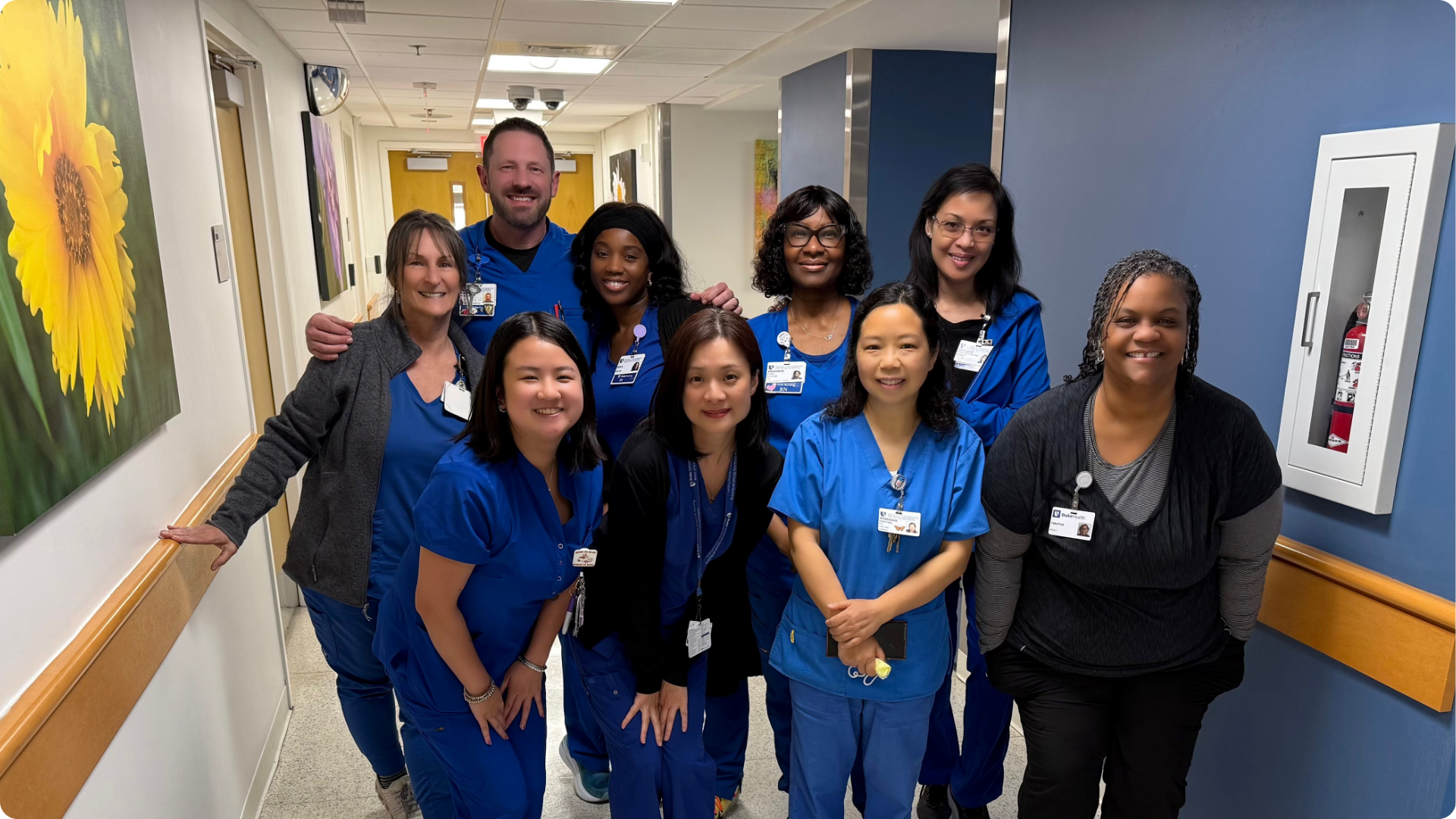 Group of nine smiling clinical research or healthcare staff members posing in a brightly lit hallway. Most are wearing blue scrubs or ID badges, indicating a professional medical or research setting. Framed artwork, including a large yellow flower, decorates the walls, and a fire extinguisher is housed in a cabinet nearby.