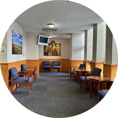 Waiting room with blue cushioned chairs and wooden armrests arranged along both walls. The room features large vertical windows on the right letting in natural light, framed with light-colored walls and wooden paneling. A few small plants sit by the windows. There is a wall-mounted TV screen and two framed nature photographs on the far wall, and a coat rack stands on the left near the entrance.