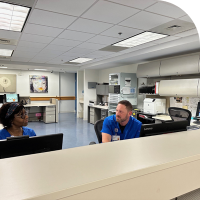 Two clinical staff members in blue scrubs talking behind a nurses' station desk in a well-lit office area. The workspace includes computers, printers, file cabinets, and wall-mounted storage units. Bright ceiling lights and modern equipment create a clean, professional atmosphere.