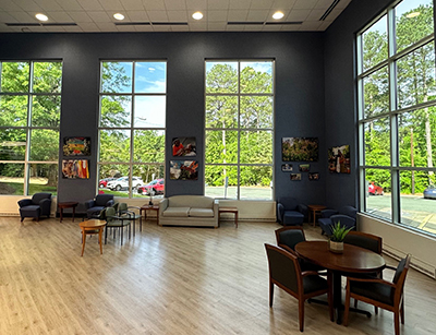 Bright, spacious lobby with floor-to-ceiling windows, light wood floors, modern seating clusters, round meeting table, and colorful artwork on navy walls.